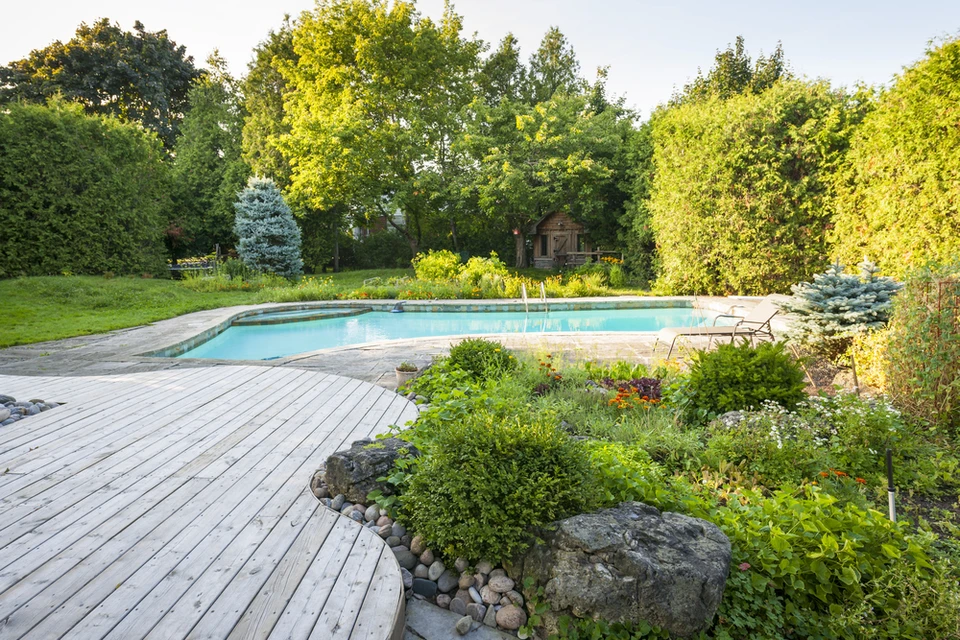 Jardin paysager avec piscine rectangulaire à débordement, terrasse en lames de bois composite, aménagement végétal avec massifs plantés, rochers décoratifs et galets